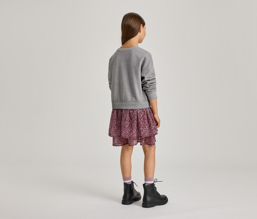 Dziewczynka od tyłu, ubrana w szarą bluzę, warstwową spódnicę w kwiaty, skarpetki w paski i czarne sznurowane botki.
