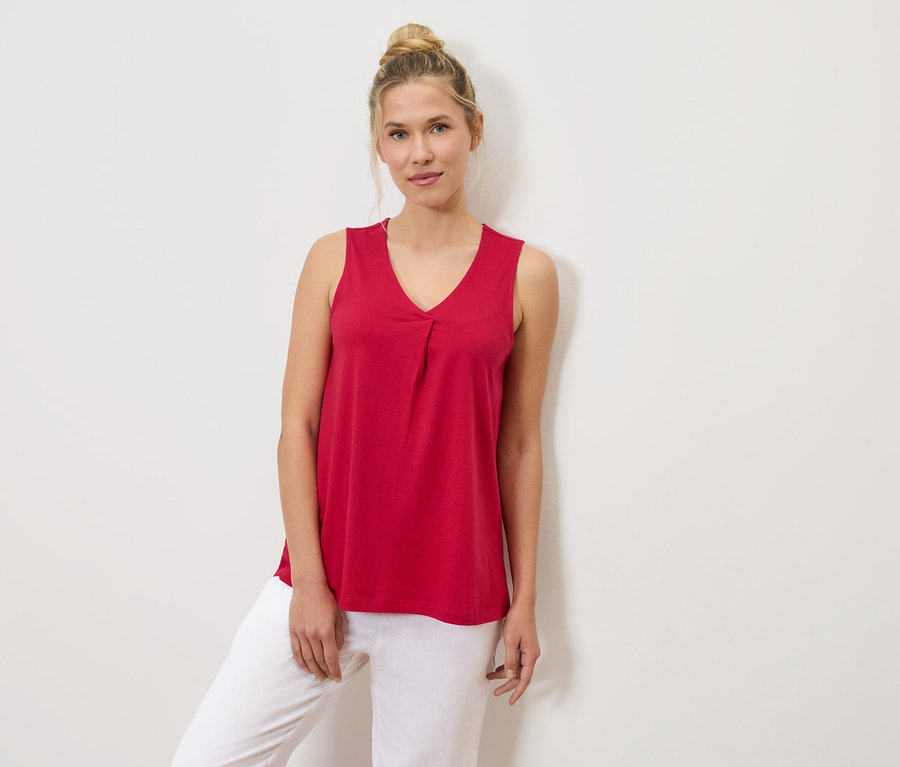 Kobieta z kokiem na głowie ma na sobie czerwony podkoszulek i białe spodnie, opierając się o białą ścianę.