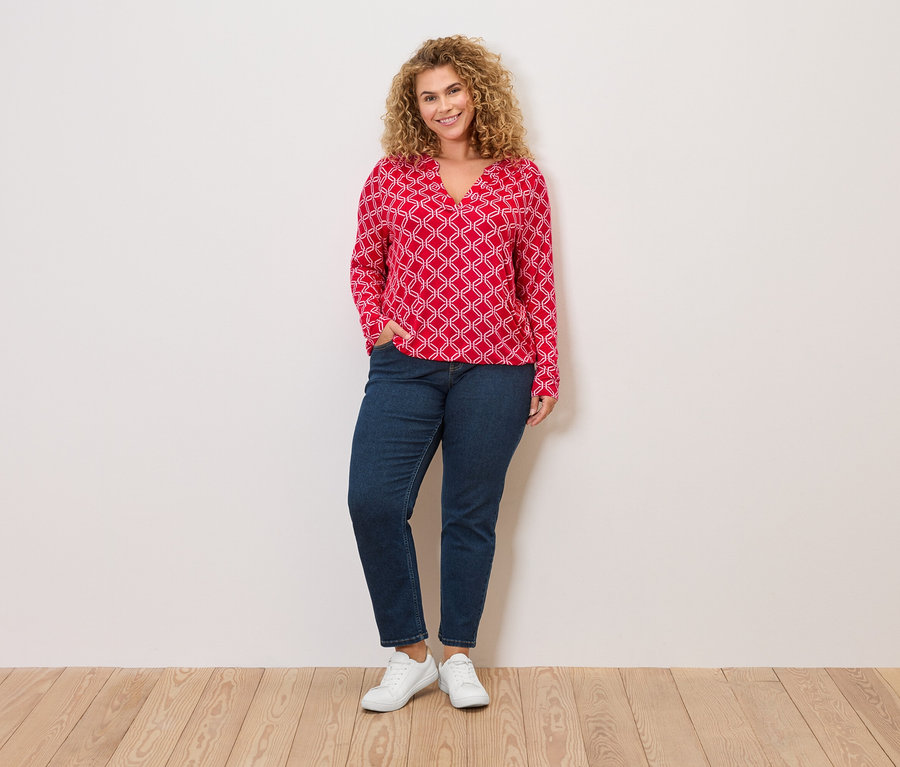 Kobieta o kręconych blond włosach, w czerwonej bluzce z białym wzorem, dżinsach i białych trampkach stoi na tle ściany.