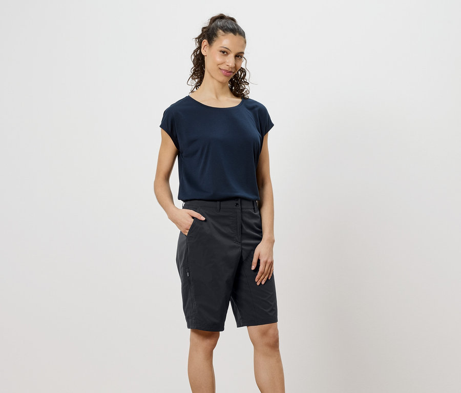 Kobieta z kręconymi, brązowymi włosami ma na sobie granatowy T-shirt i szare szorty, z ręką w kieszeni.