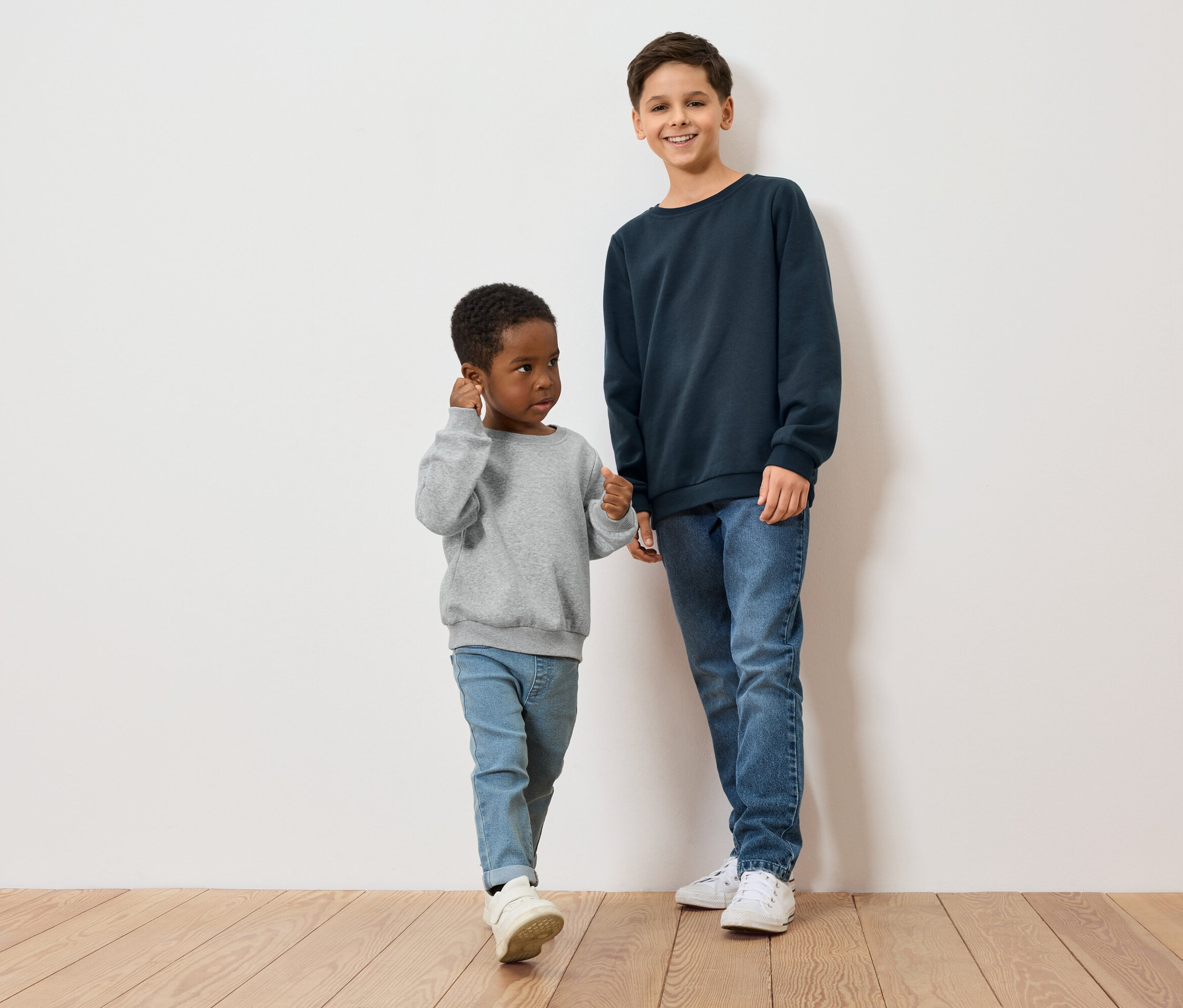 Dwóch chłopców, jeden z ciemną skórą w szarej bluzie, drugi z jasną skórą w niebieskiej bluzie, stoi na drewnianej podłodze przed białą ścianą.