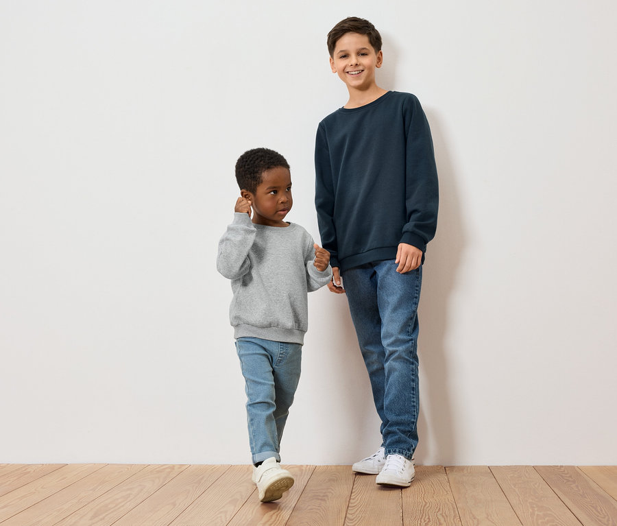 Dwóch chłopców, jeden z ciemną skórą w szarej bluzie, drugi z jasną skórą w niebieskiej bluzie, stoi na drewnianej podłodze przed białą ścianą.