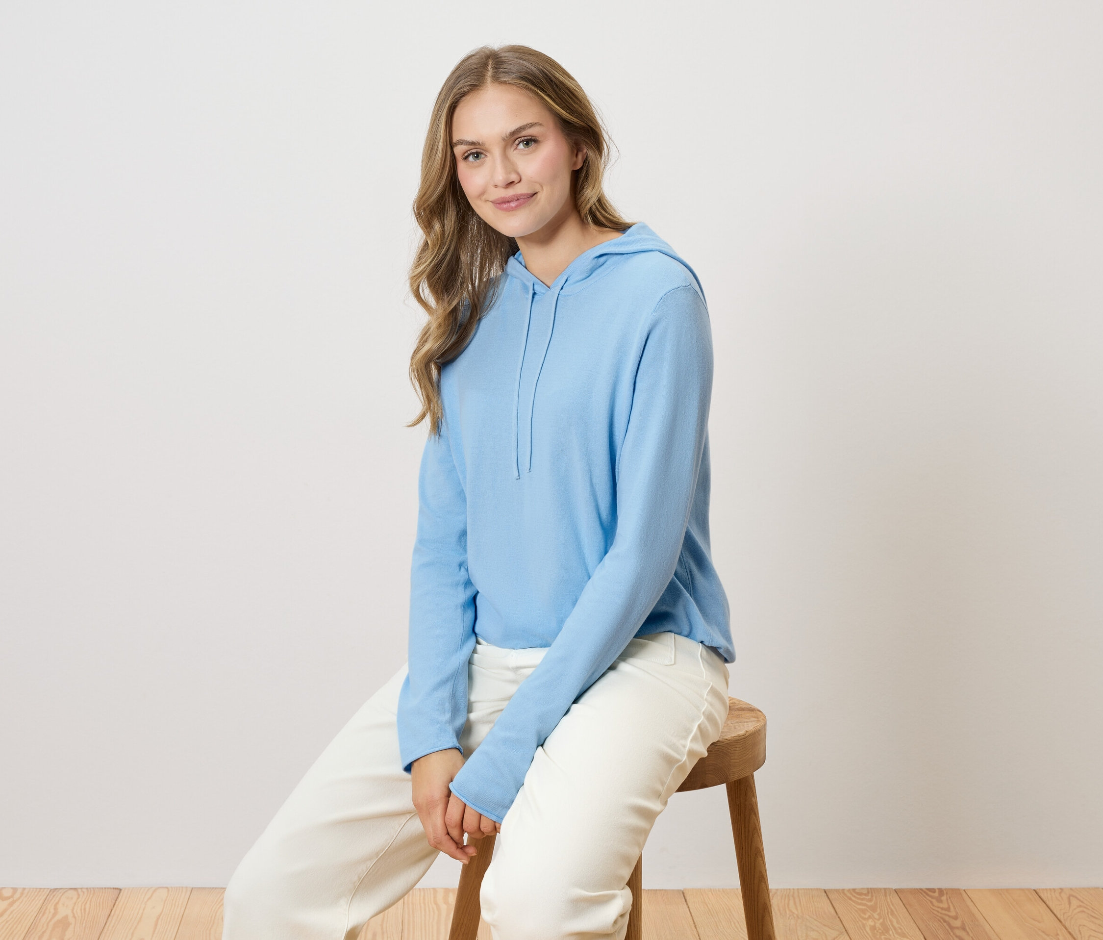 Kobieta z długimi blond włosami siedzi na drewnianym krześle, ubrana w jasnoniebieską bluzę z kapturem i białe spodnie.