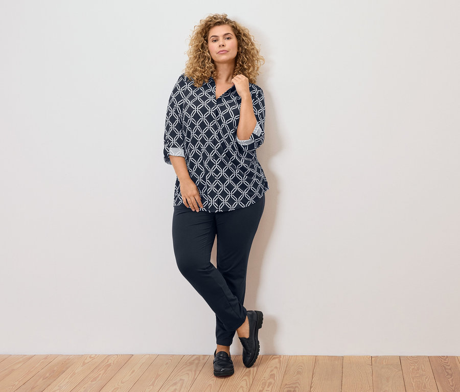 Kobieta o kręconych blond włosach, ubrana w bluzkę w niebiesko-biały wzór, ciemne spodnie i czarne buty, opiera się o ścianę.