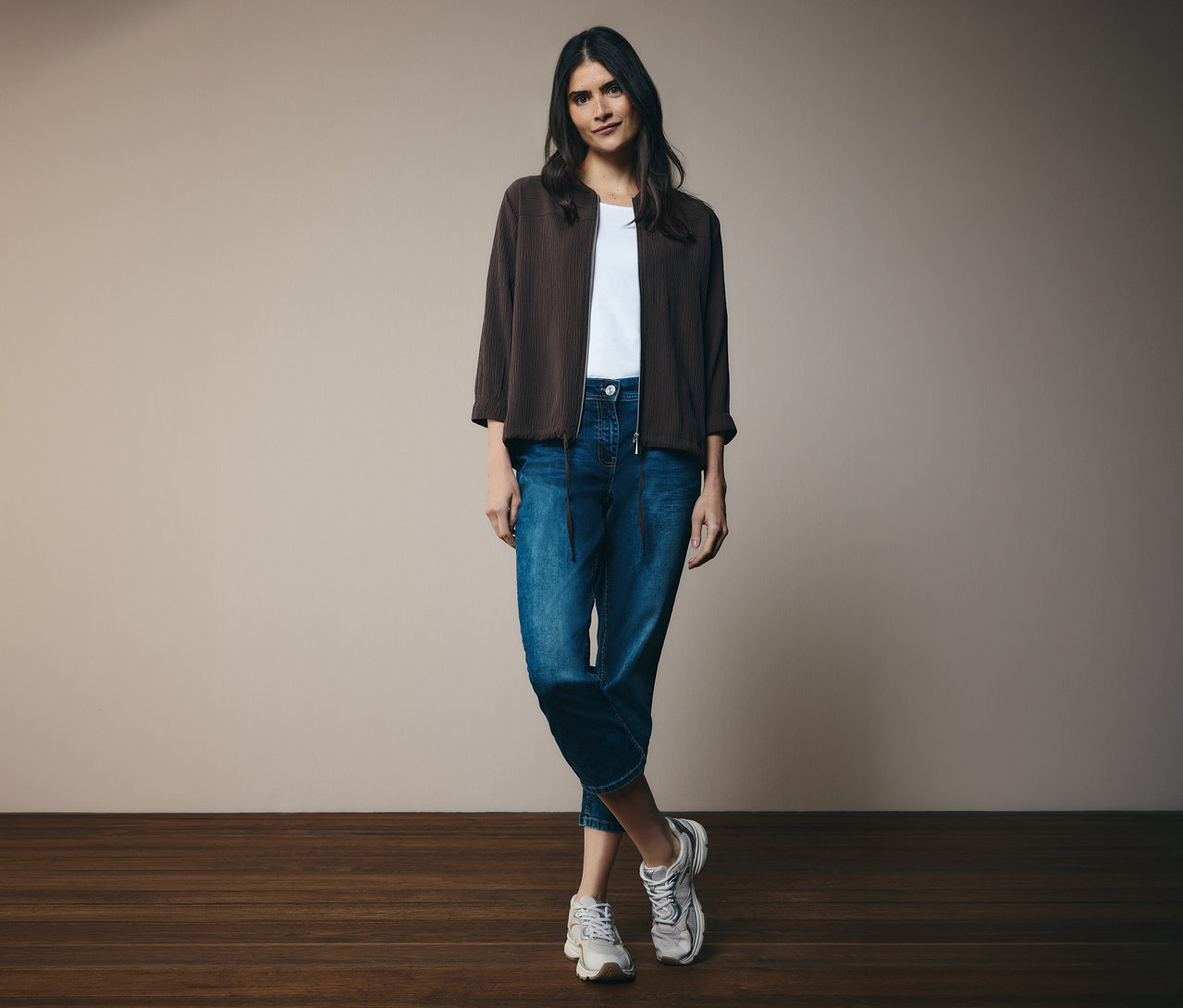 Kobieta w białym T-shircie, dżinsach i brązowej kurtce bomberce CECIL. Stoi na tle ściany.