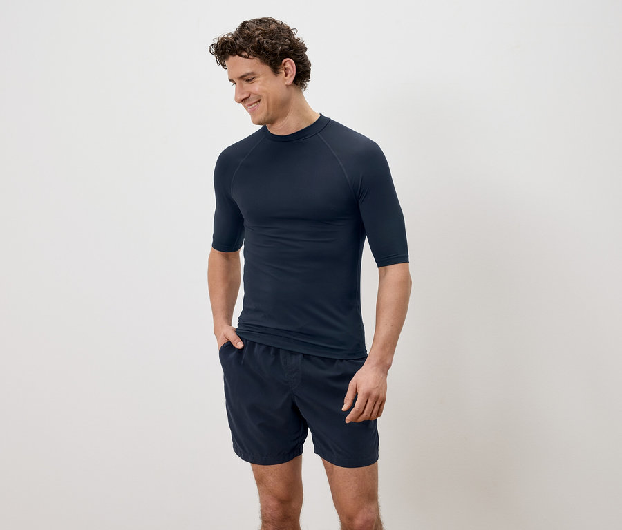 Mężczyzna z kręconymi włosami w granatowej koszulce z krótkim rękawem i szortach na tle zwykłego tła.