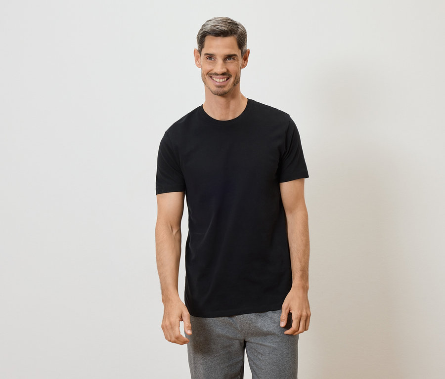 Uśmiechnięty mężczyzna z siwymi włosami w czarnym T-shircie i szarych spodniach na białym tle.