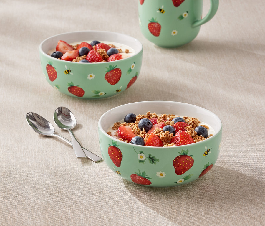 Dwie miski wypełnione jogurtem, truskawkami i jagodami oraz dzbanek we wzór truskawek stoją na stole.