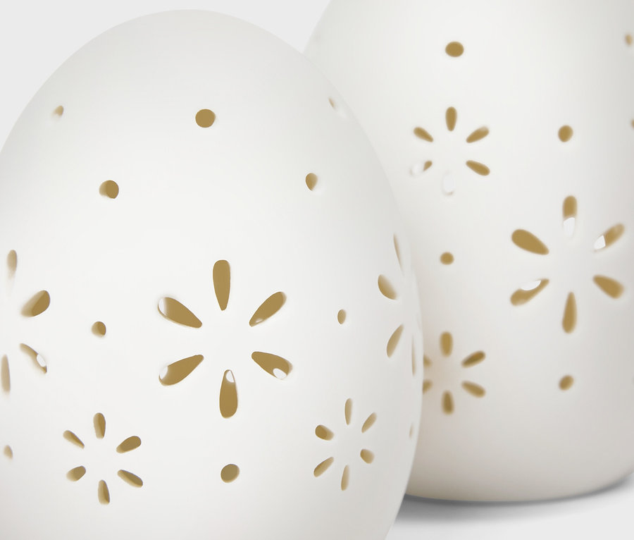 Dwie białe, ceramiczne dekoracje w kształcie jajka z wyciętymi wzorami kwiatów i kropek.