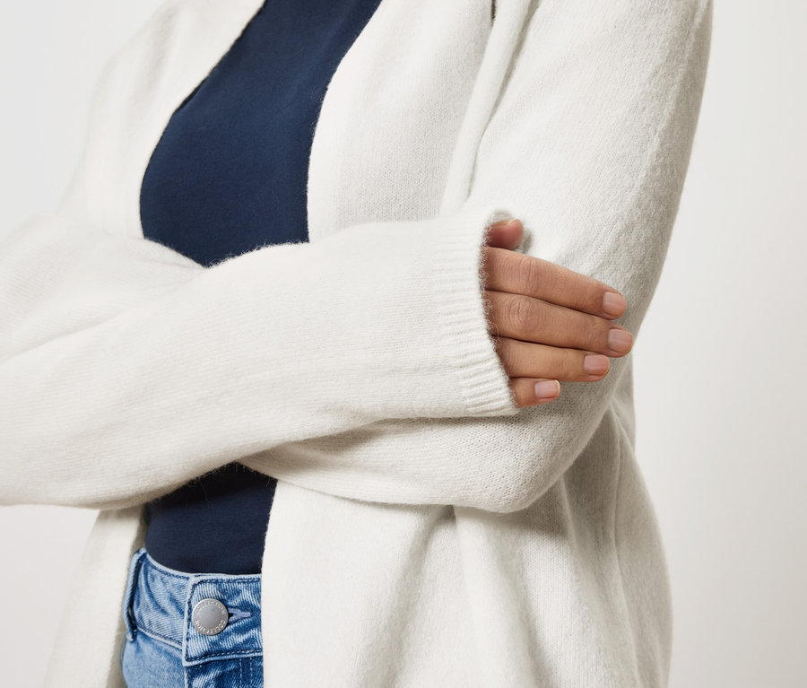 Kobieta ze skrzyżowanymi ramionami ma na sobie długi, biały kardigan na granatową bluzkę i niebieskie dżinsy.