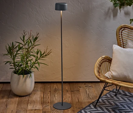 Lampa podłogowa stoi obok rośliny doniczkowej i wiklinowego krzesła z poduszką na wzorzystym dywanie.