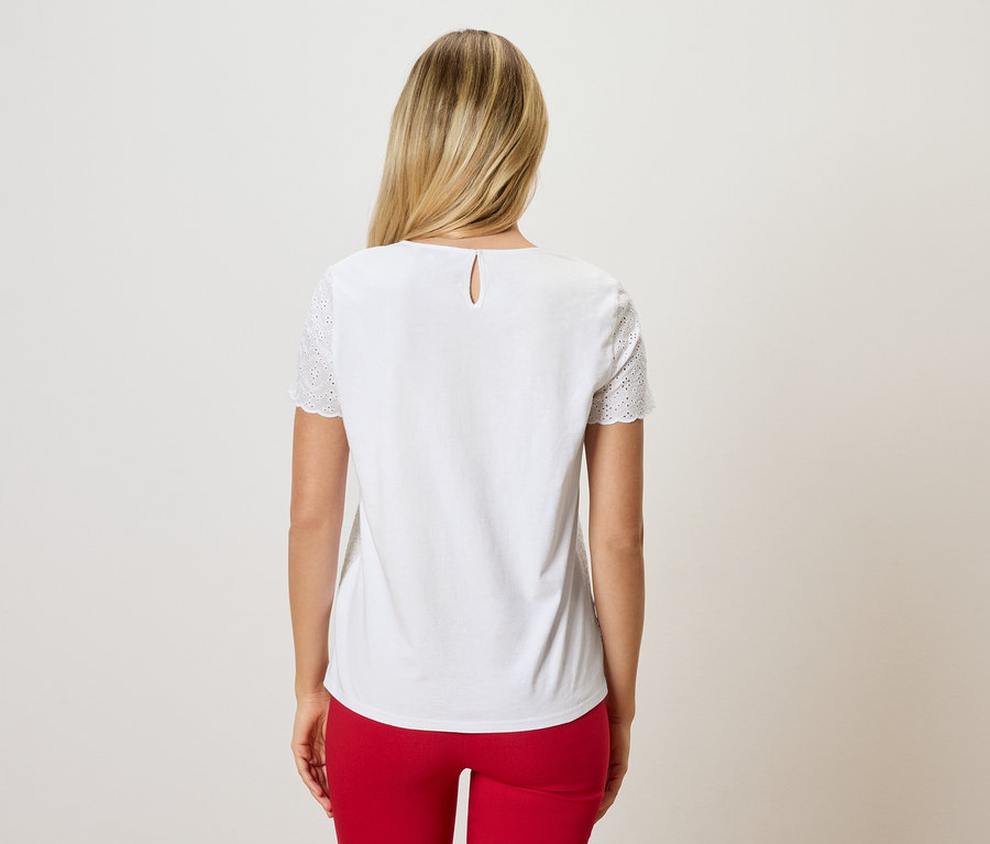 Widok z tyłu kobiety z blond włosami, ubranej w białą bluzkę z haftem dziurkowanym na rękawach i czerwone spodnie.