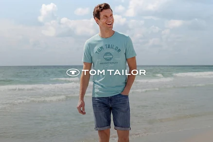 Uśmiechnięty mężczyzna w jasnoniebieskiej koszulce z nadrukiem i dżinsowych szortach stoi na plaży z morzem w tle. Logo Tom Tailor.