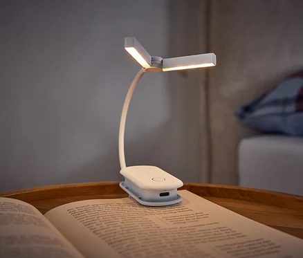 Biała lampka do książek z klipsem oświetla otwartą książkę leżącą na drewnianej tacy. W tle widać łóżko z poduszką w kratę.