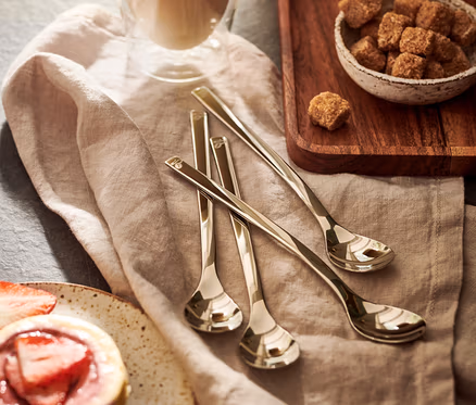 Szklanka latte macchiato, obok miska z brązowym cukrem i babeczka z truskawkami. Na serwetce leży zestaw 4 łyżeczek do latte macchiato.