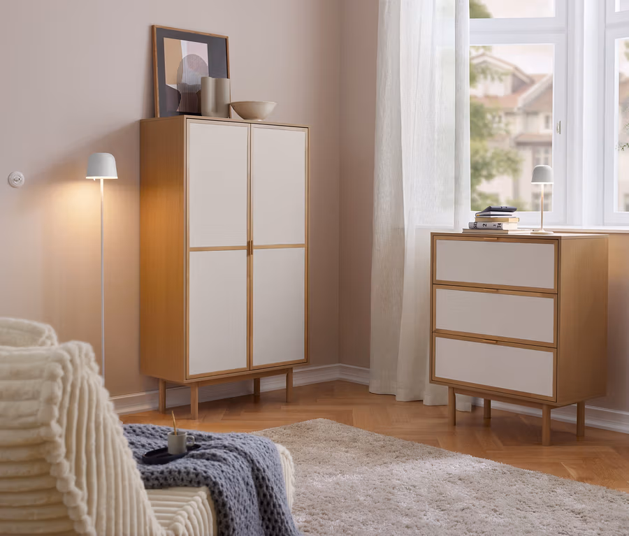 Jasny salon z drewnianą szafką Cabinetschrank z 2 drzwiami i komodą z 3 szufladami z jasnymi frontami. Na komodzie stoi akumulatorowa lampa stołowa LED Eglo „Mannera”. Po lewej sofa, po prawej okno z zasłonami.