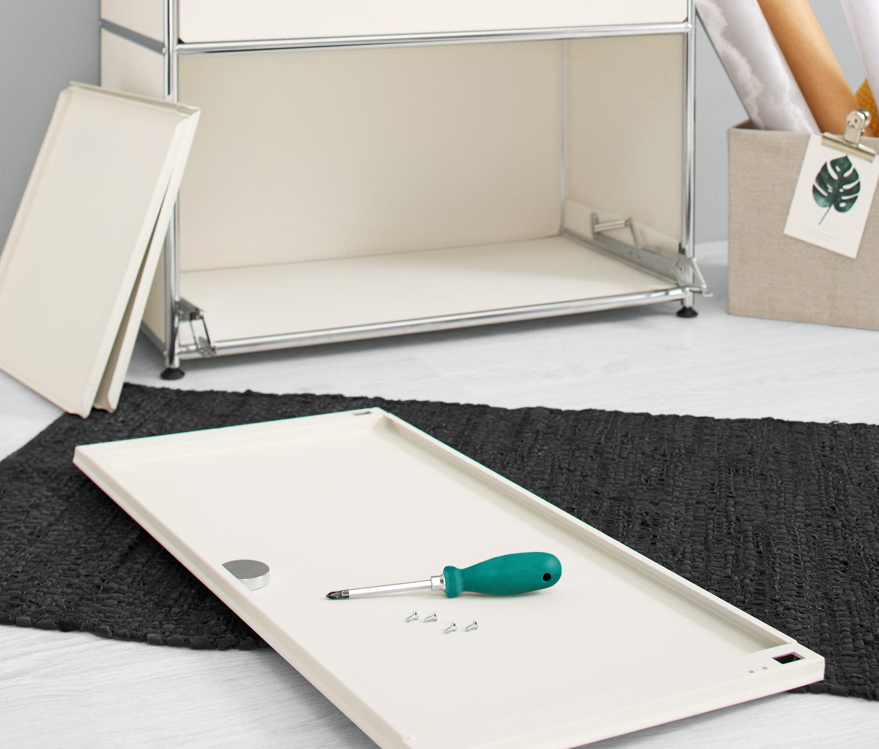 Na podłodze leży biała dodatkowa klapa metalowa, zestaw »CN3«, ze śrubokrętem i śrubami obok komody.