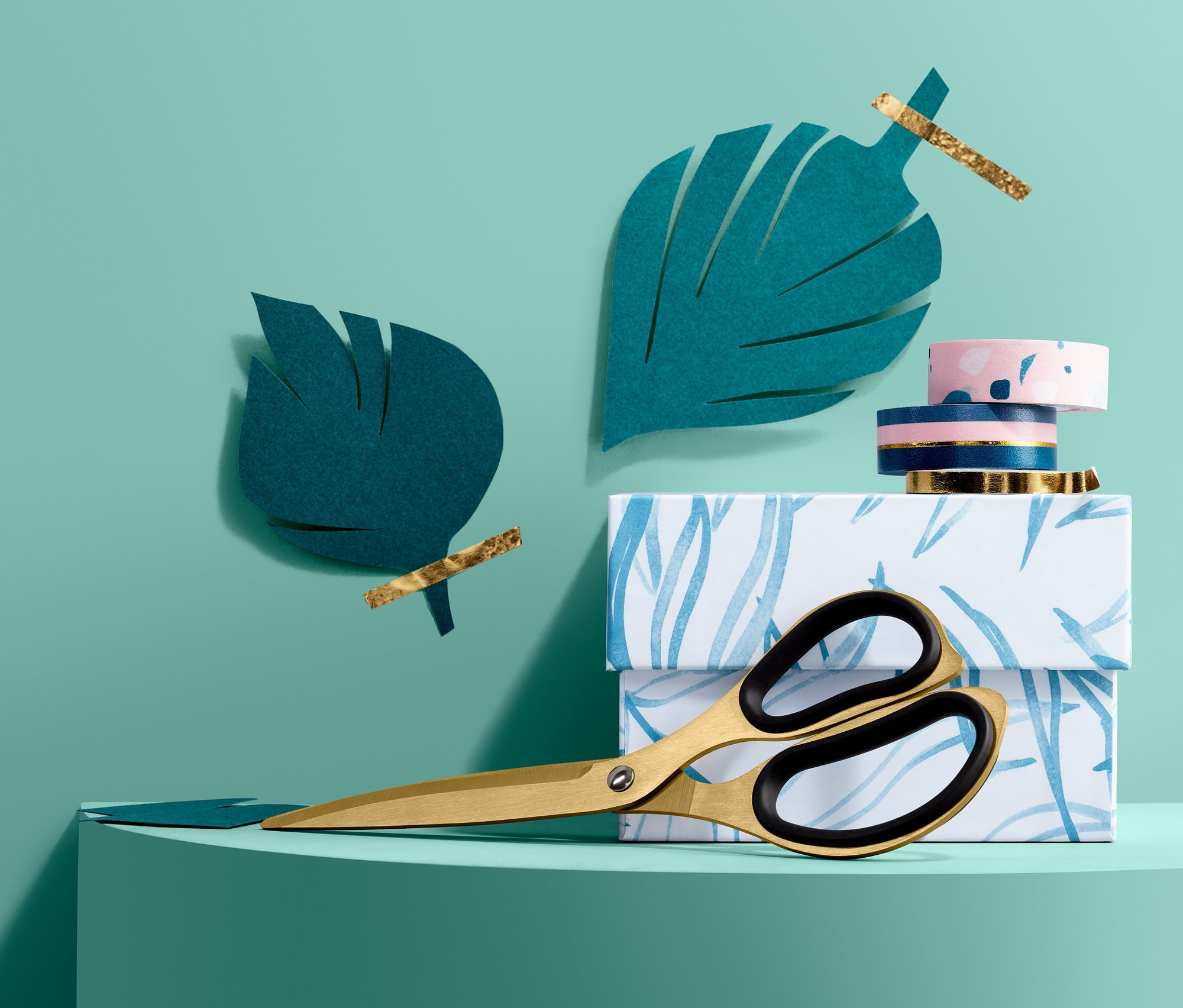 Złote nożyczki biurowe leżą na pudełku z niebieskim wzorem liści obok papierowych liści.