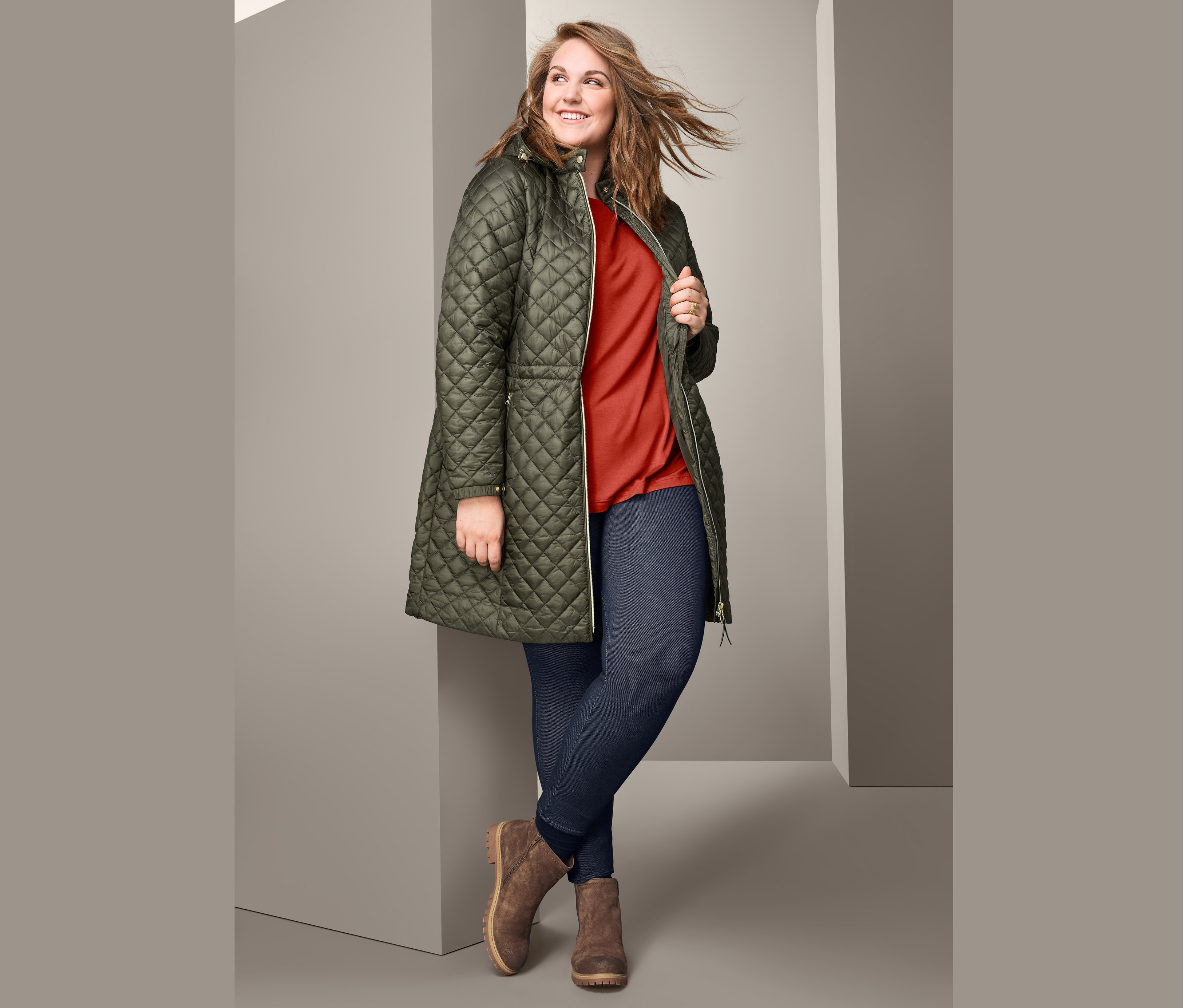 Kobieta pozuje w damskim płaszczu pikowanym z kapturem, damskiej bluzce oversize, legginsach z podszewką stylizowanych na jeansy i damskich botkach z weluru.