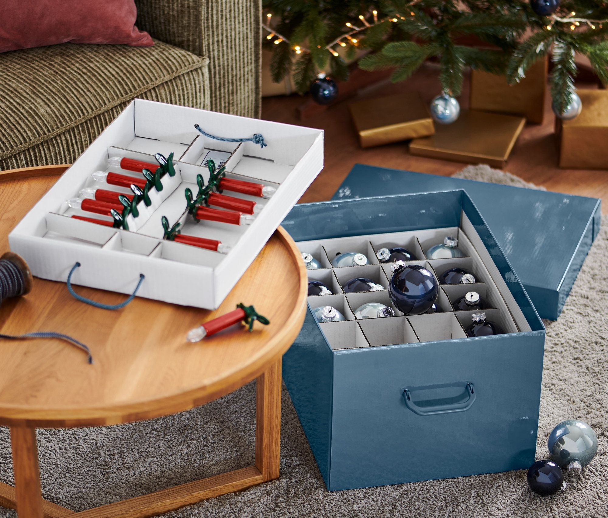 Otwarte pudełko na ozdoby bożonarodzeniowe z czerwonymi świecami i niebieskimi bombkami obok choinki.