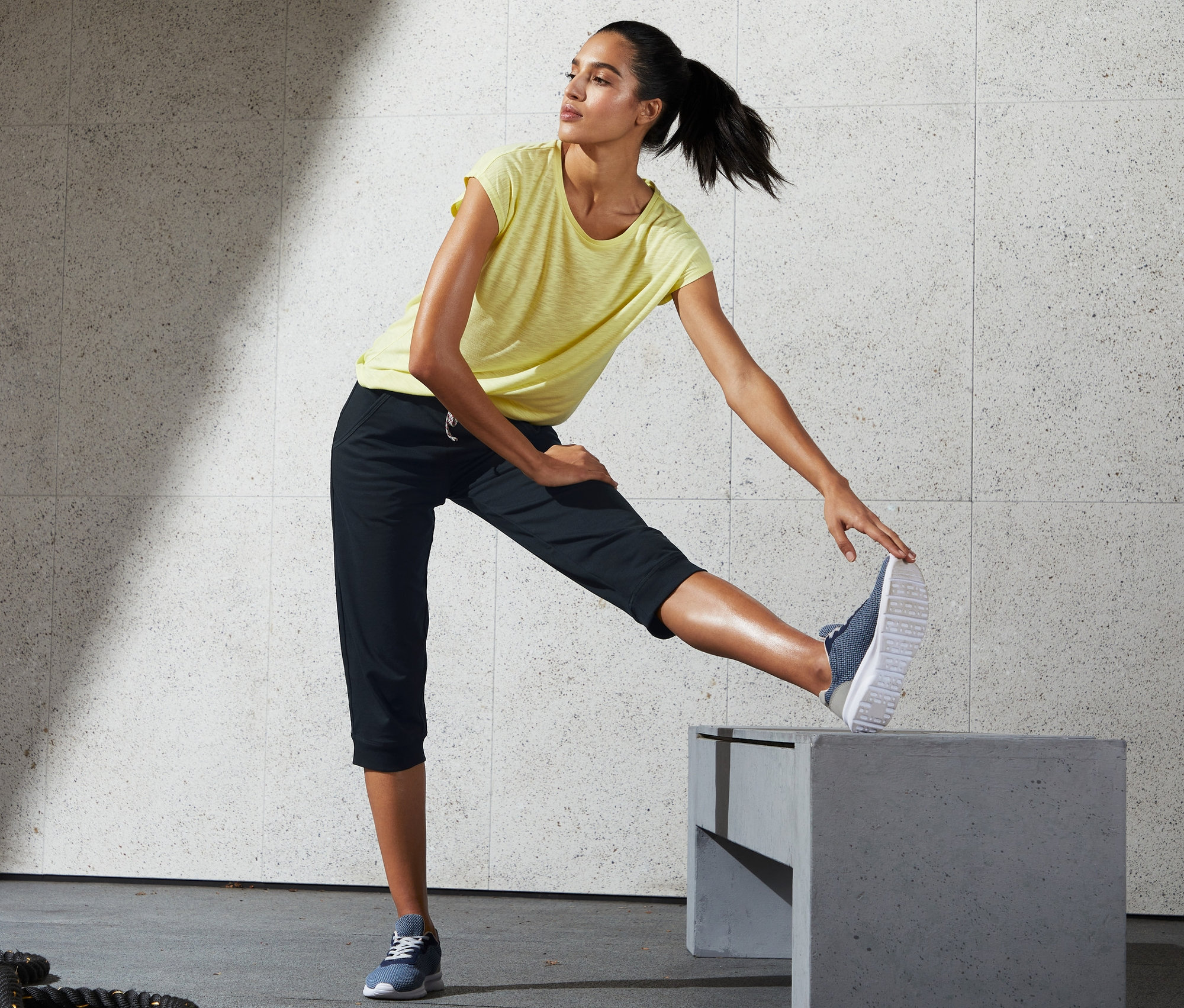 Kobieta w żółtej koszulce i spodniach sportowych o długości 3/4 rozciąga się na szarym bloku.