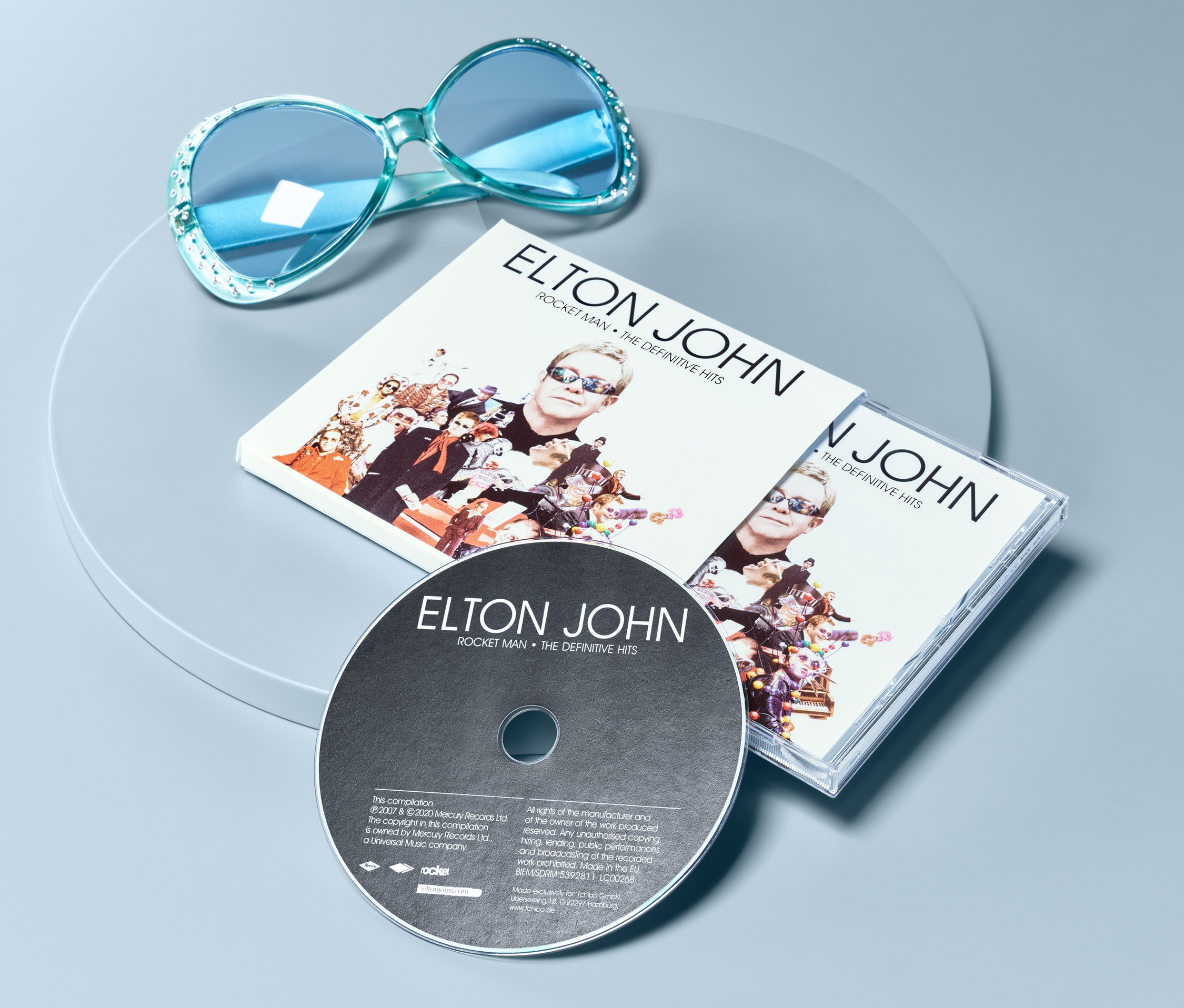 Niebieskie okulary przeciwsłoneczne i płyta CD Elton John – »Rocket Man: The Definitive Hits« na szarym tle.