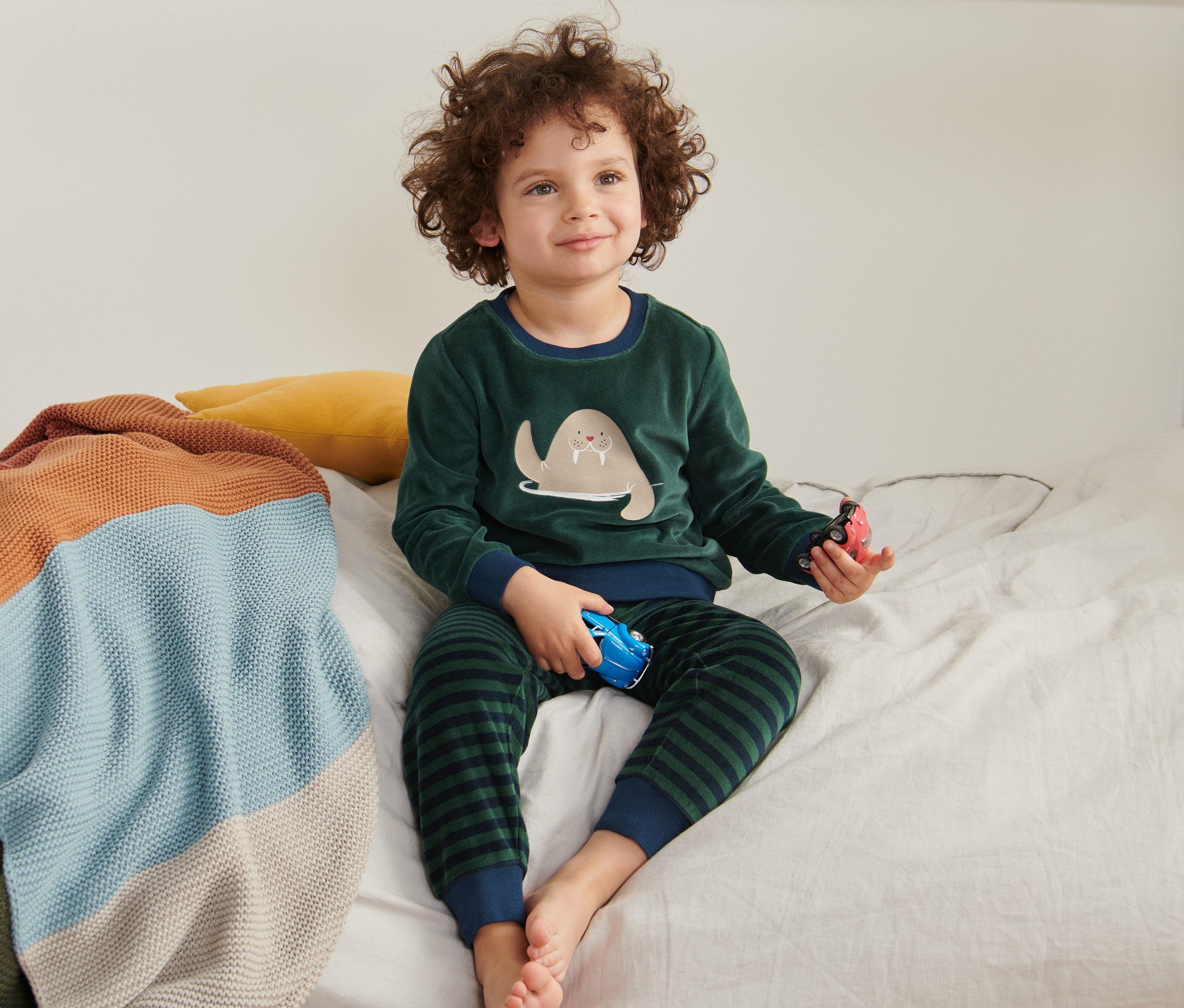 Chłopiec siedzi na łóżku w welurowej piżamie dziecięcej z morsem i bawi się autami.