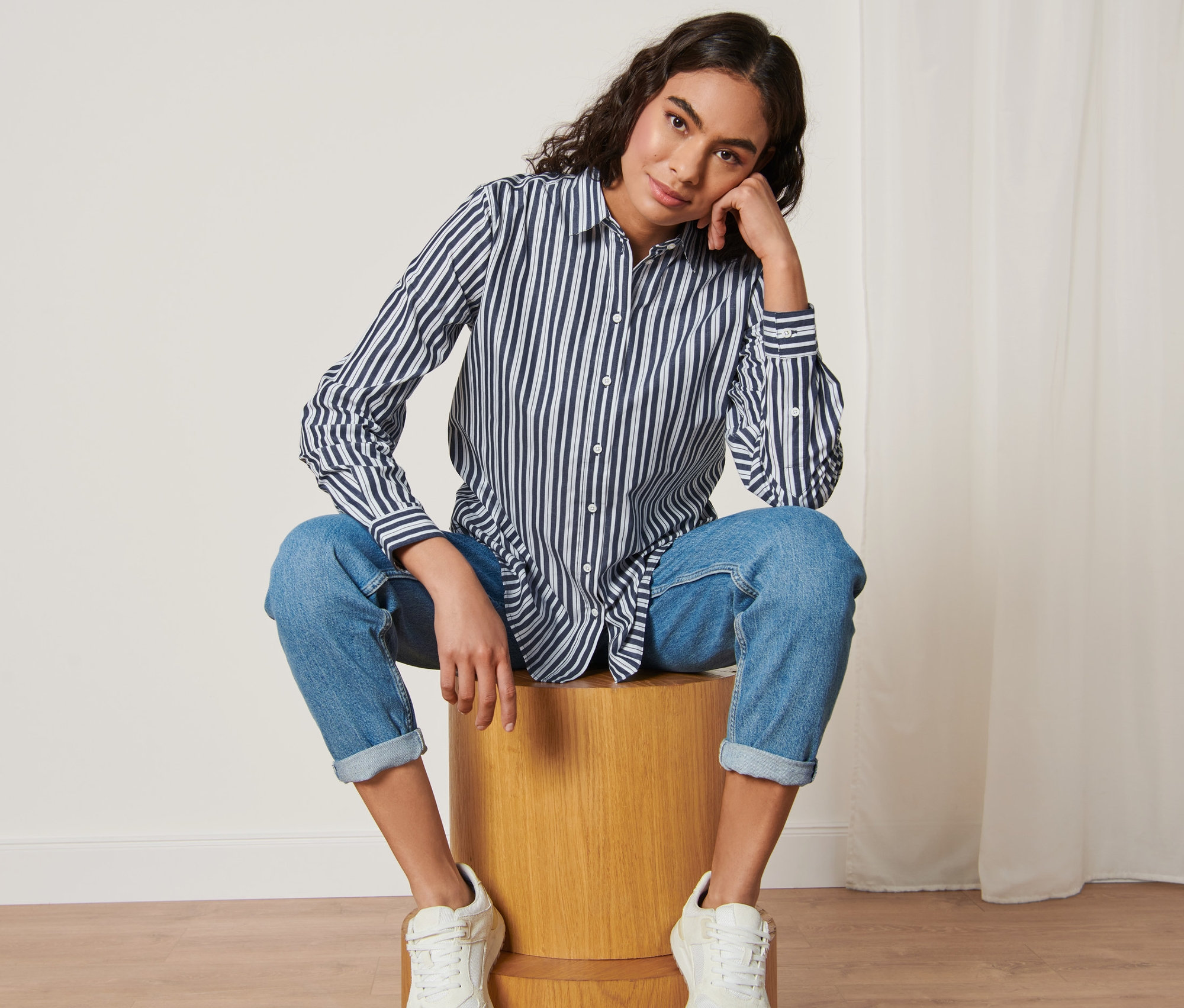 Kobieta siedzi na drewnianym stołku i ma na sobie Koszula z popeliny w paski.