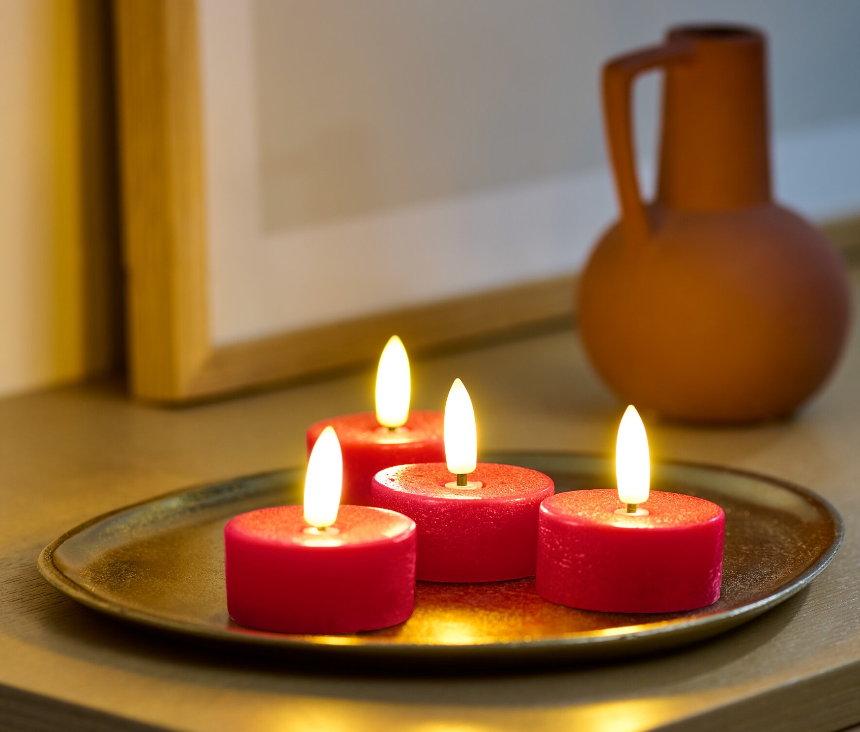 Cztery czerwone świeczki typu tealight LED z prawdziwego wosku na złotej tacy na szafce.