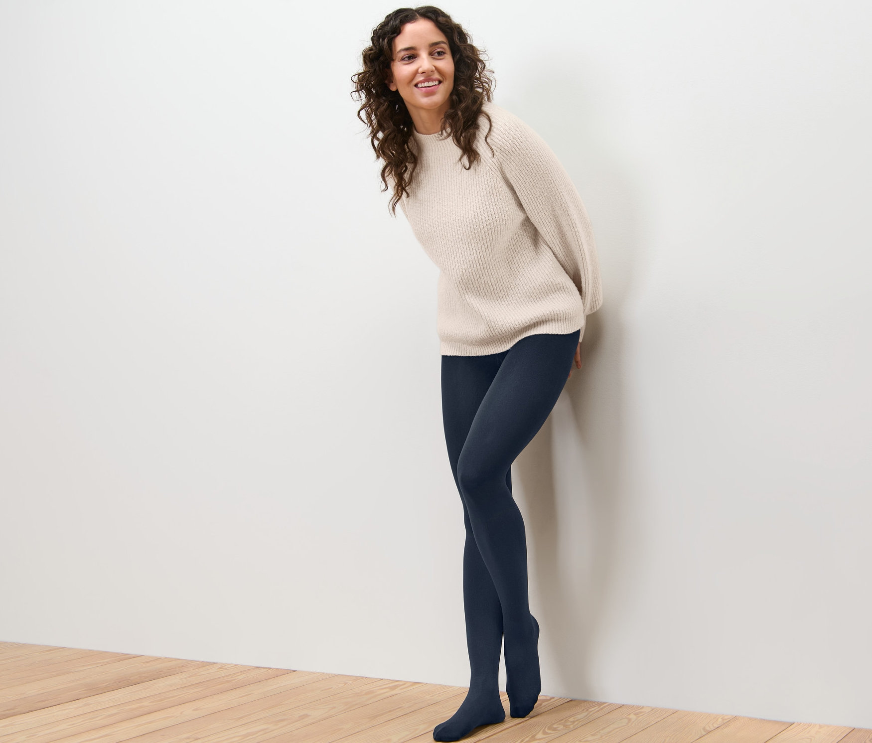 Uśmiechnięta kobieta oparta o ścianę ma na sobie rajstopy termoaktywne i sweter.