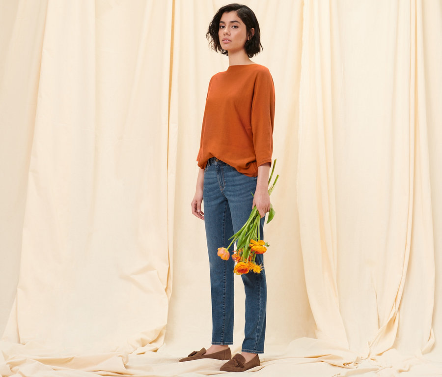 Kobieta pozuje na tle materiału, trzymając tulipany. Ma na sobie pomarańczowy sweter z delikatnej dzianiny, jeansy slim »Fit Emma« w kolorze mid blue denim i półbuty ze skóry welurowej.