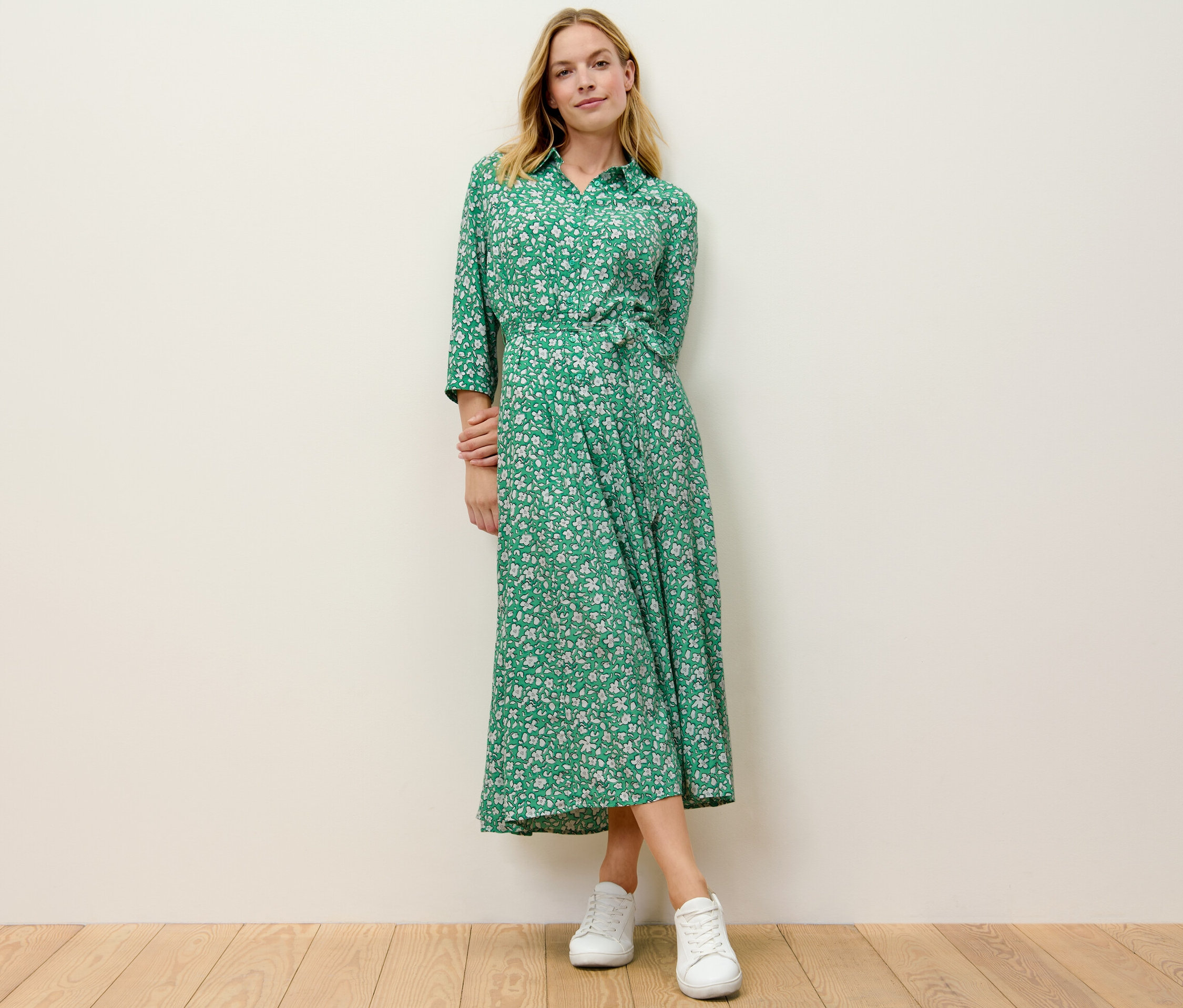 Blondynka stoi w zielonej wzorzystej sukience koszulowej.