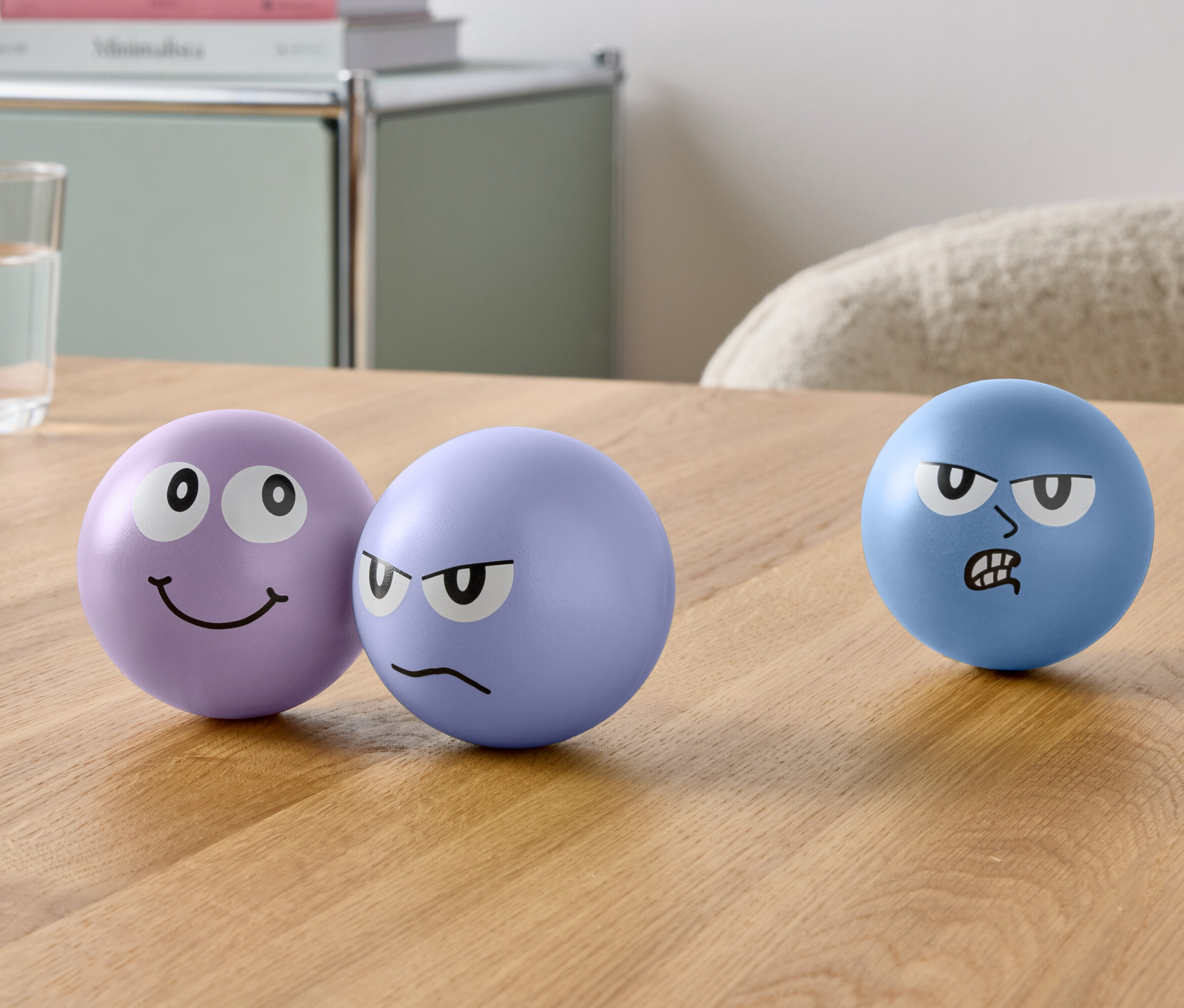 Na stole stoją trzy kule z narysowanymi twarzami i szklanka wody.