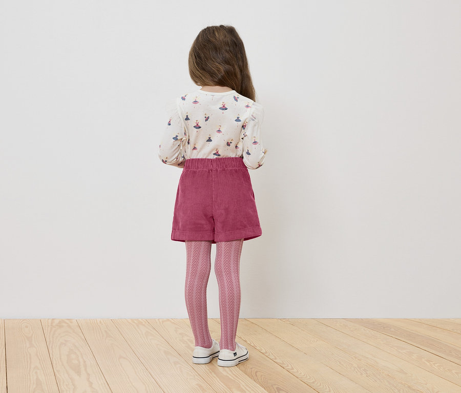 Dziewczynka stoi tyłem, ma na sobie bluzkę z baletnicami, różowe szorty i dziergane rajstopy.