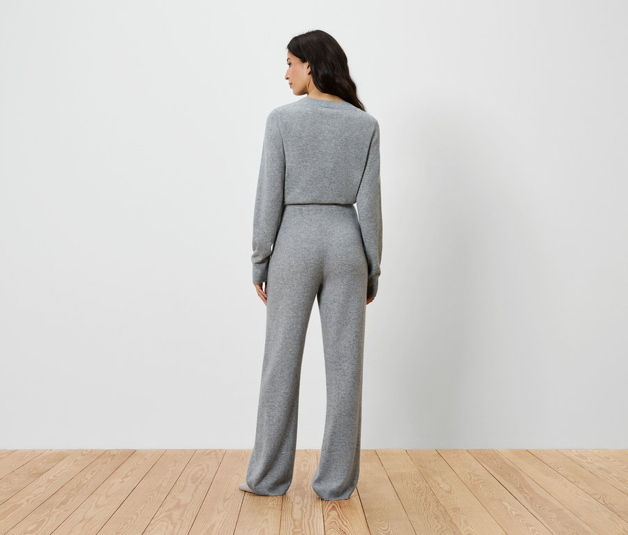 Kobieta stoi tyłem do aparatu, ubrana w szary sweter z długim rękawem i szerokie spodnie.