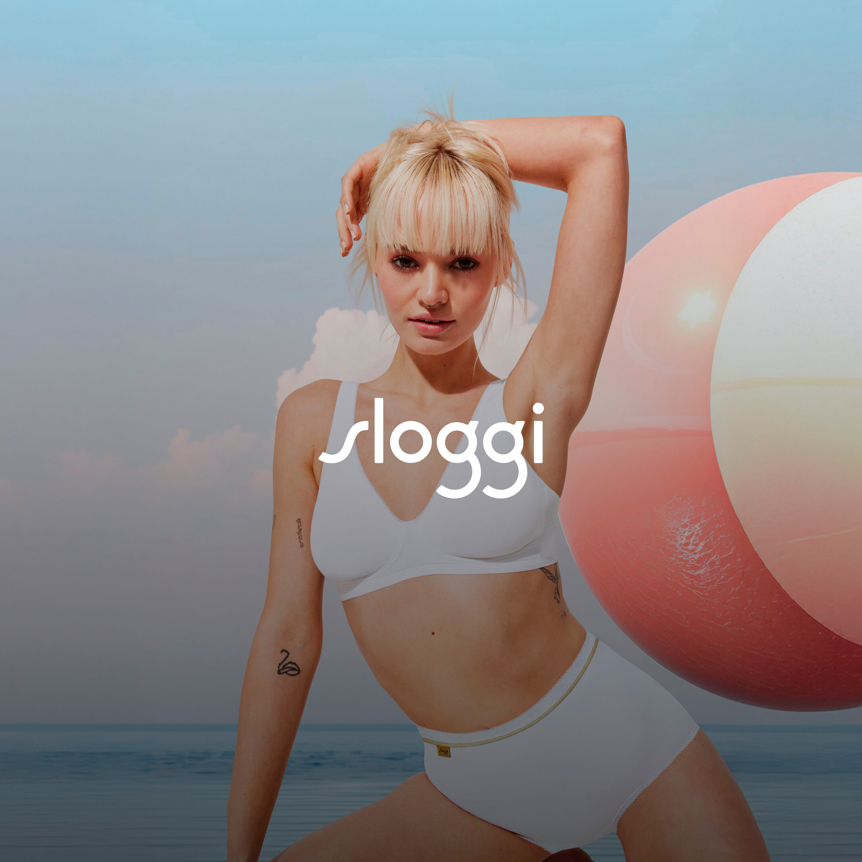 Blondwłosa kobieta w białej bieliźnie pozuje z ręką nad głową obok piłki plażowej.