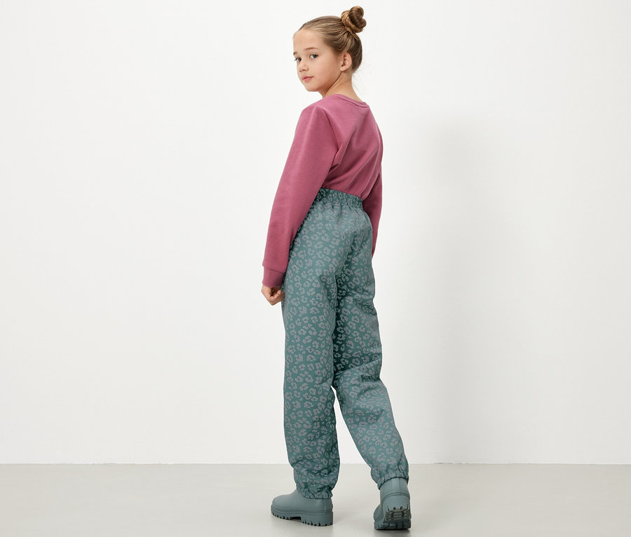 Dziewczynka w bluzie i dziecięcych spodniach przeciwdeszczowych z elementami odblaskowymi, widziana od tyłu.