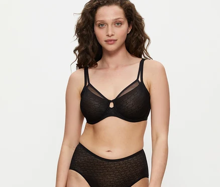 Kobieta z kręconymi brązowymi włosami ma na sobie czarny biustonosz Triumph Signature Sheer Minimizer i pasujące figi z wysokim stanem, patrzy w kamerę.