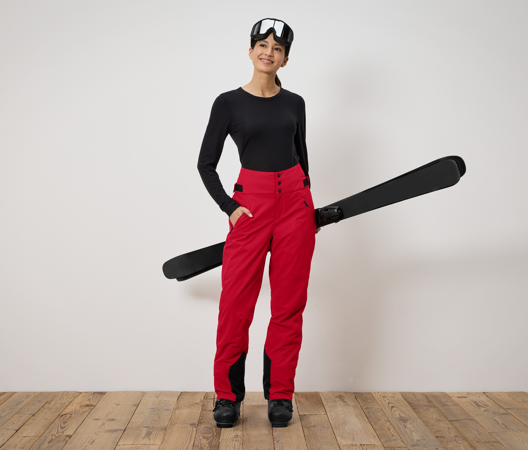 Kobieta stoi i trzyma narty. Ma na sobie czarną bluzkę z długim rękawem, czerwone spodnie i gogle narciarskie.