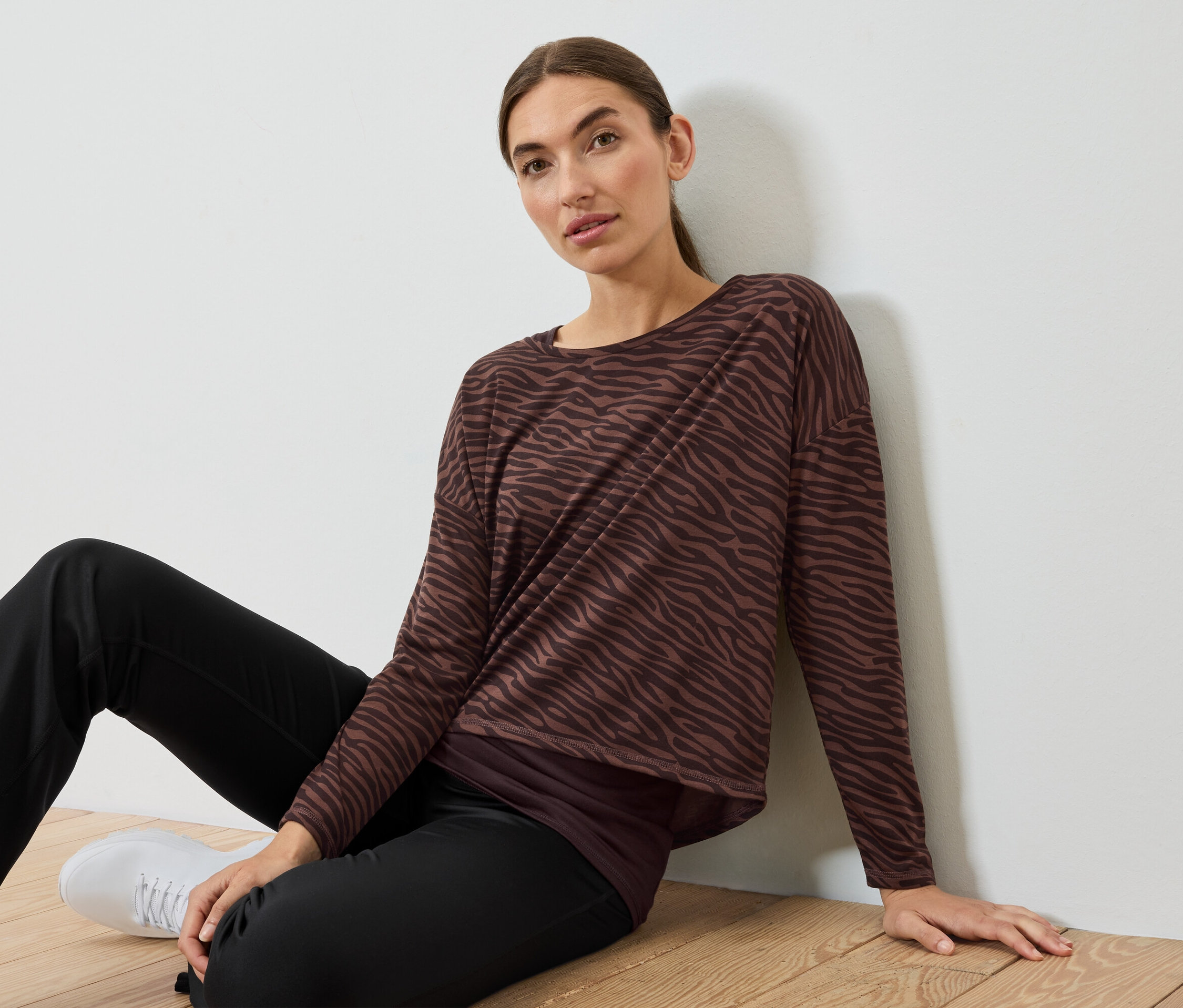 Kobieta siedzi na podłodze w brązowo-czarnym topie w zebrę i czarnych spodniach.