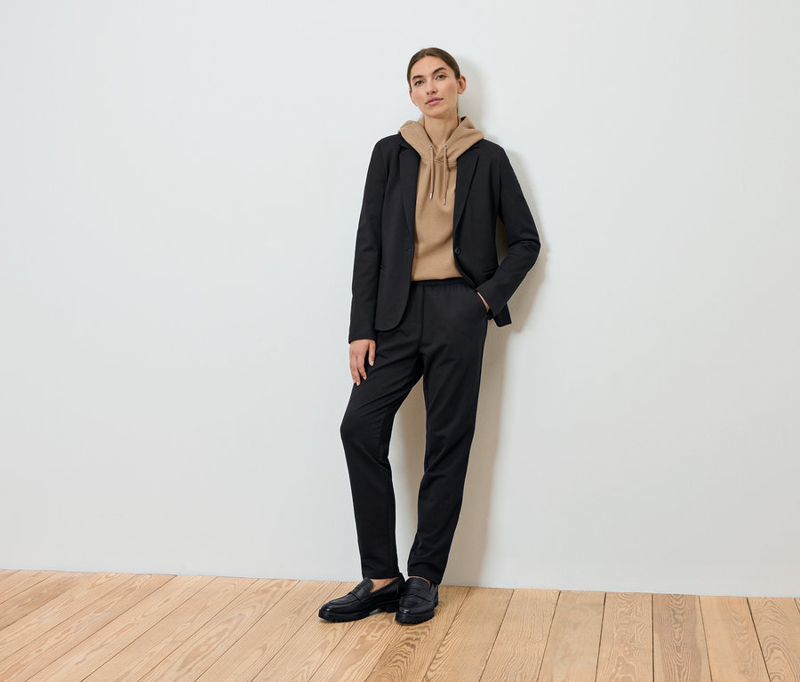 Kobieta pozuje w czarnej marynarce, brązowej bluzie z kapturem i czarnych spodniach.