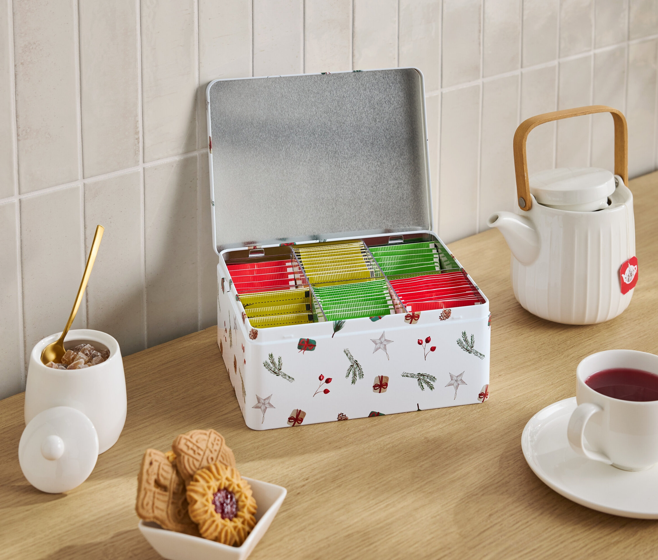 Otwarta metalowa puszka z motywami świątecznymi wypełniona torebkami herbaty stoi obok białego czajnika, filiżanki herbaty, cukierniczki z cukrem i miski z ciastkami.