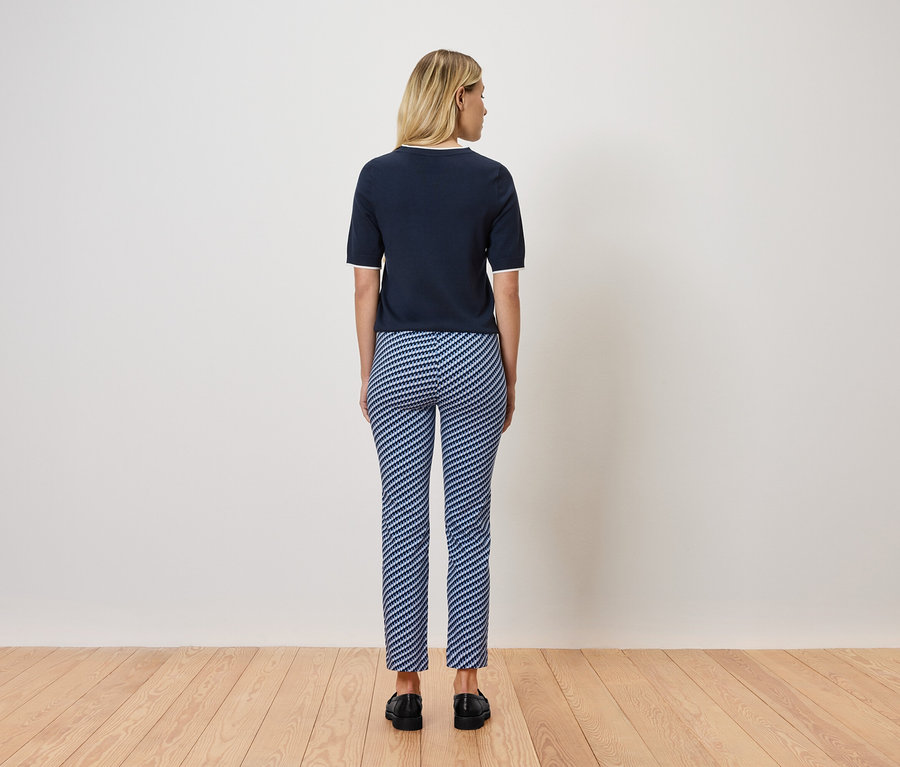 Blondynka stoi tyłem do kamery, ubrana w granatowy top z krótkim rękawem i niebiesko-białe spodnie w paski. Stoi na drewnianej podłodze.