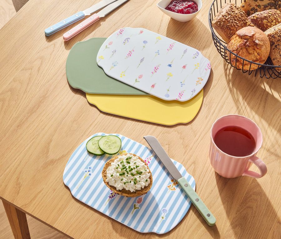 Na drewnianym stole leżą chleby w koszyku, dżem, noże i deski do krojenia w różnych kolorach.