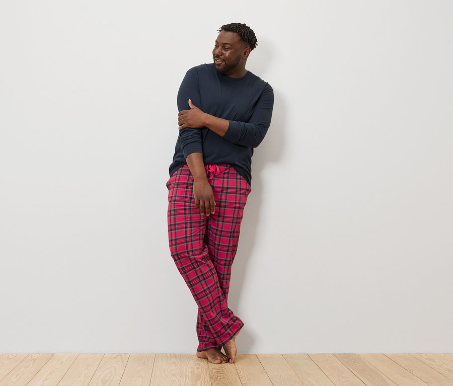 Mężczyzna oparty o ścianę ma na sobie granatową koszulę z długim rękawem i czerwoną piżamę w kratę.