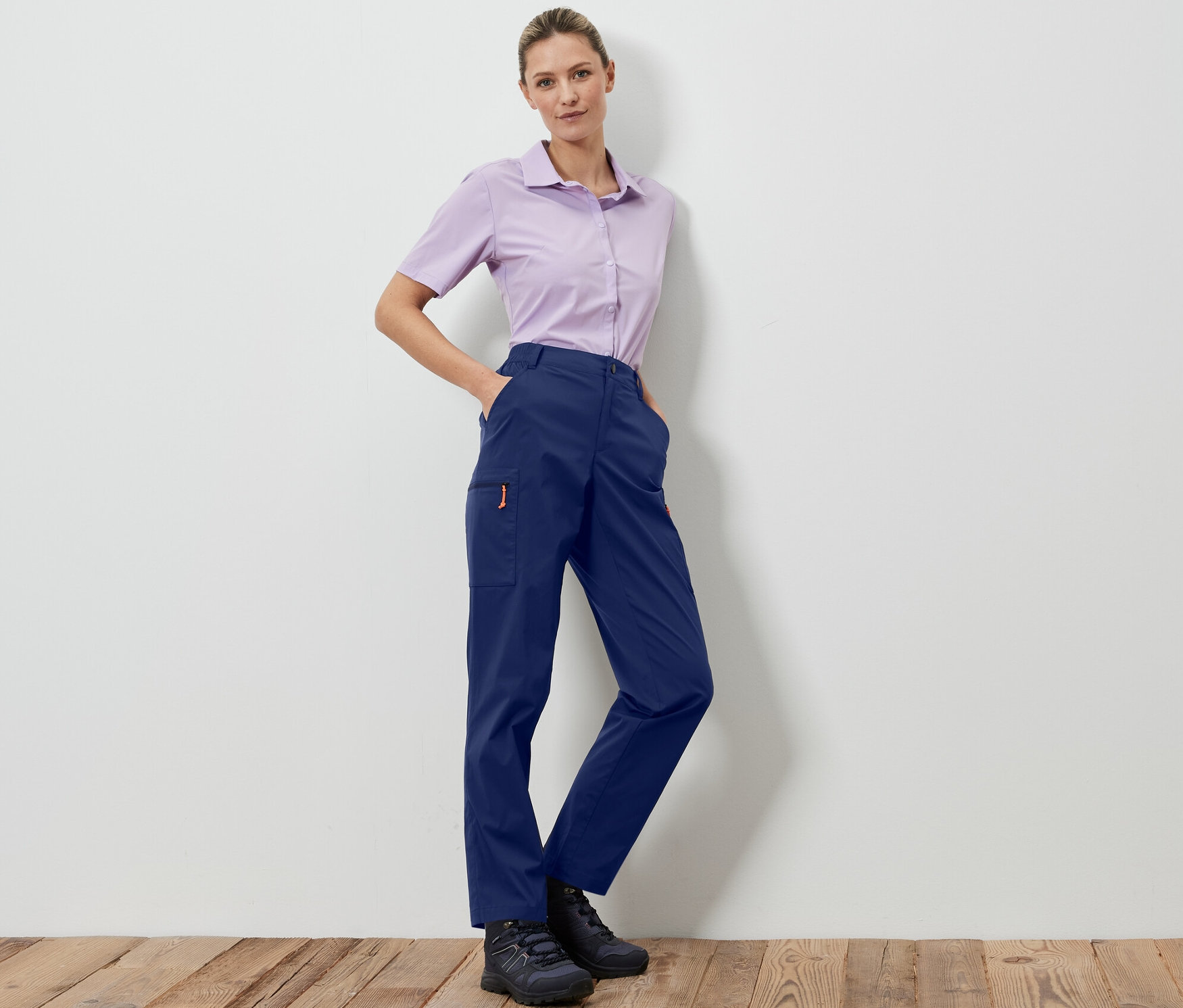 Kobieta pozuje w granatowych spodniach outdoorowych z możliwością spakowania i jasno fioletowej koszuli.