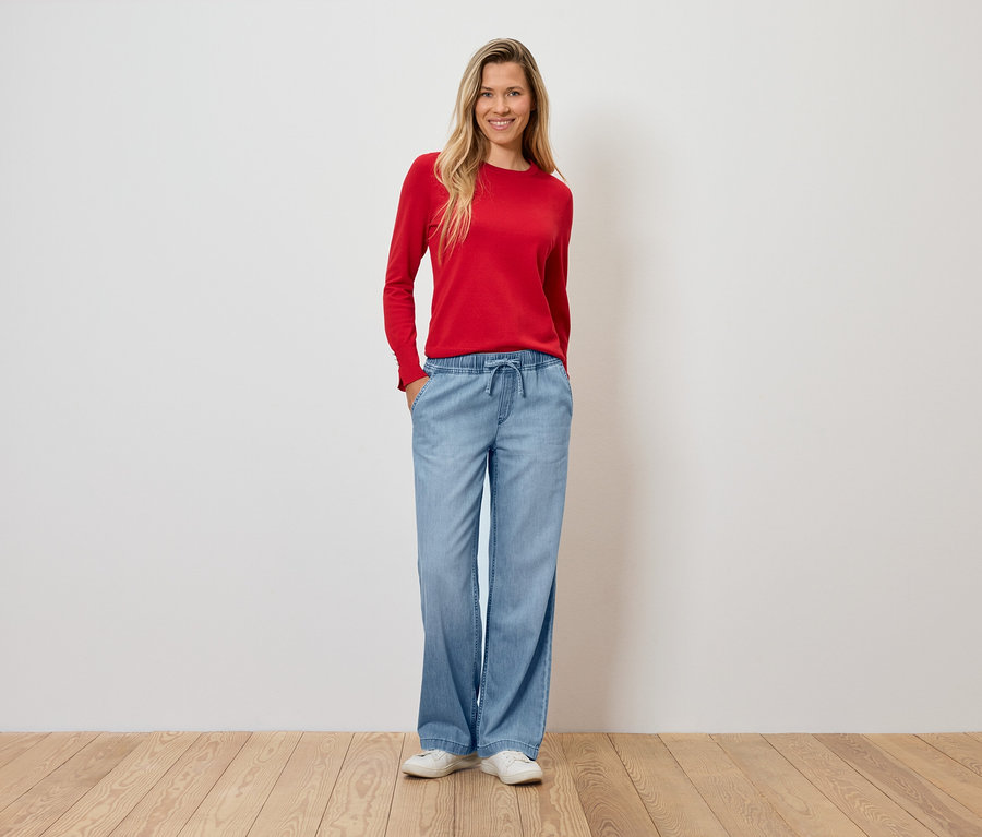 Kobieta w czerwonym swetrze i szerokich, jasnoniebieskich spodniach stoi na drewnianej podłodze.