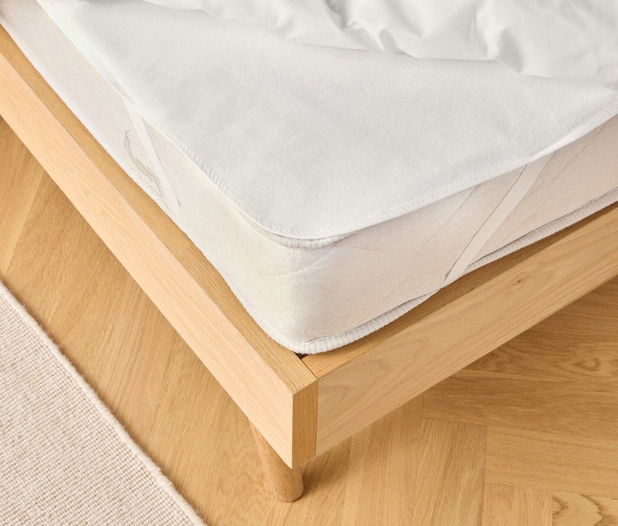 Widok na jasną drewnianą ramę łóżka z białym materacem i białą kołdrą. Częściowo widoczny jest beżowy dywan.