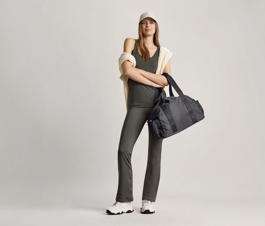 Kobieta pozuje w oliwkowym topie i czarnej torbie sportowej.