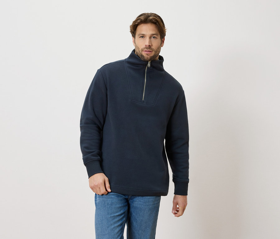 Mężczyzna nosi granatowy sweter z zamkiem błyskawicznym i niebieskie dżinsy.